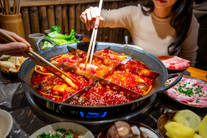 Dos personas usando palillos para sumergir carne en un caldero dividido lleno de caldo de olla caliente picante y burbujeante