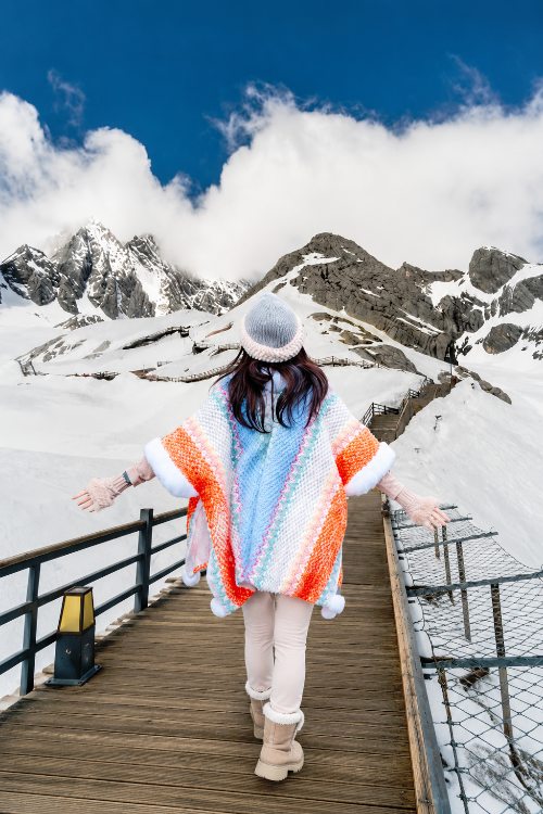 Turista con poncho y gorro camina por un sendero rodeado de montañas nevadas y cielo azul.